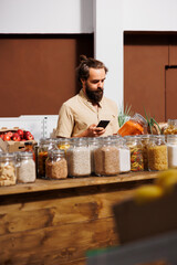 Man in zero waste store analyzing pantry products, using smartphone to make sure they are freshly harvested. Vegan customer thoroughly checking local supermarket food items are preservatives free - obrazy, fototapety, plakaty