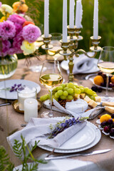 Romantic sunset wedding table decor for a couple with vintage chandelier, burning candles, snacks, white wine, fruits, ranunculi flowers. Summer vibes, field or meadow, outdoors event