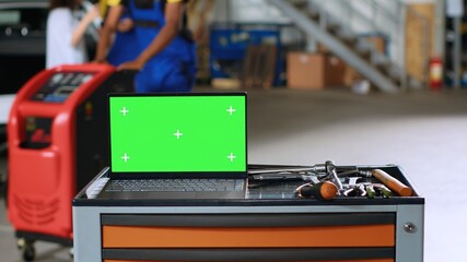 Green screen laptop placed on working bench in busy garage next to professional tools while...