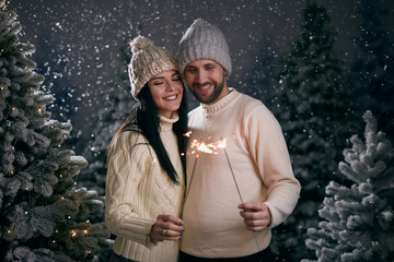 guy and girl in white sweaters with sparklers in a snowy forest, winter and christmas concept