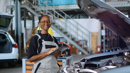 Portrait of happy african american mechanic doing car maintenance in repair shop using tablet to keep track of operations. Expert in garage looking over checklist on device while fixing vehicle