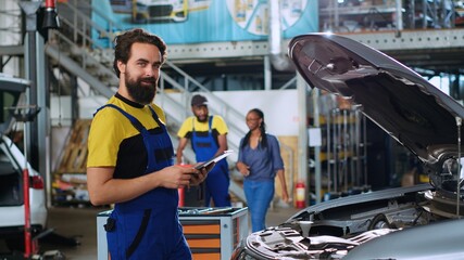 Portrait of smiling skillful mechanic in auto repair shop doing car annual checkup using tablet,...