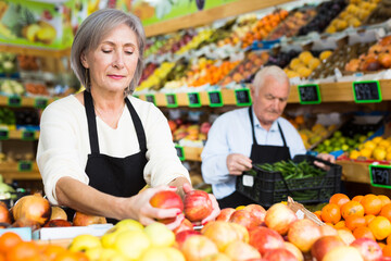 Greengrocer workers, old man and woman, doing their job in salesroom. Woman putting goods on shelves, man holding crate full of green pepper.