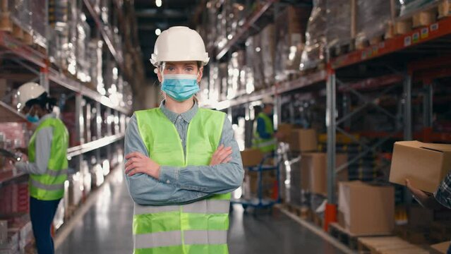 Female worker standing at storage of logistic enterprise. Woman industrial employee looking at camera at background of multi tiered racks. Lady wearing uniform and crossing arms at stock corridor.