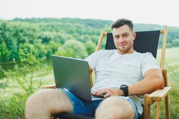 Obraz premium View of handsome guy lying on chair in nature and working with laptop, resting alone, thoughtfully looking up. A man on a trip, in the countryside. the concept of people's lifestyle