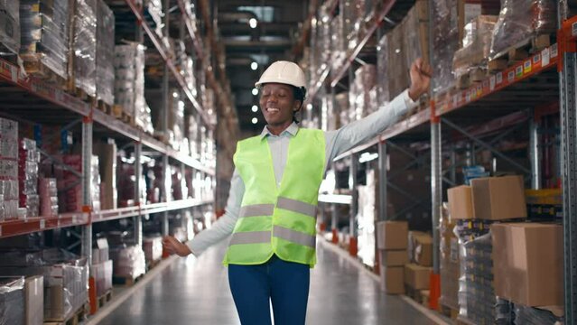 African American woman cheerfully dancing at storage building. Happy female worker having fun at warehouse. Industry employee relaxing during break. Funny woman waving hands at workplace.