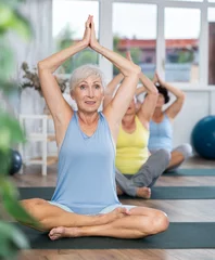Fotobehang Calm aged woman performing lotus position of yoga in fitness room with other mature attendees © JackF
