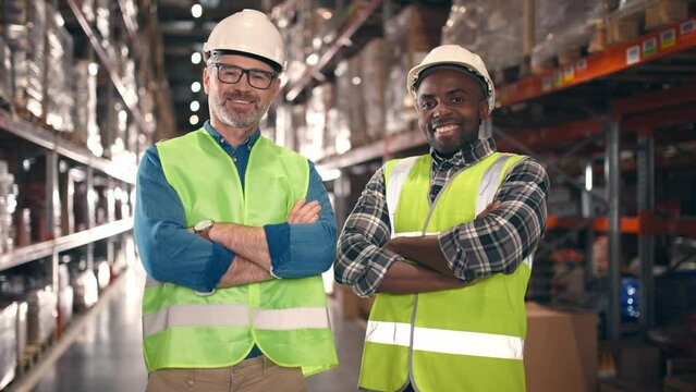 Multicultural men in protective uniform looking at each other at warehouse. Two industrial workers smiling at background of multi tiered shelves. Storage employees with arms crossed.