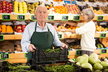 Old man supermarket worker in apron standing in salesroom with crate full of green pepper in hands....