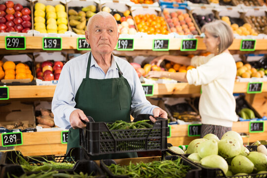 Old man supermarket worker in apron standing in salesroom with crate full of green pepper in hands. Mature woman shopping in background.