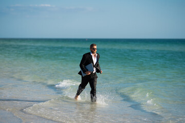 Business man in suit with laptop running in sea water. Travel tourism and business concept. Crazy male office employee with laptop running in sea. Crazy business man on summer vacation.
