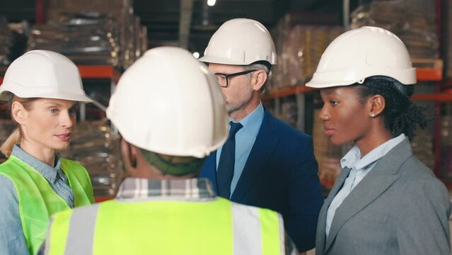 Male foreman conducting staff meeting. Industrial colleagues talking during work break. Multicultural team in reflective vests and hard hats on background of warehouse with materials.