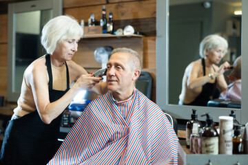 Positive experienced female barber using clippers while doing classic haircut to elderly gray-haired male client in barbershop..