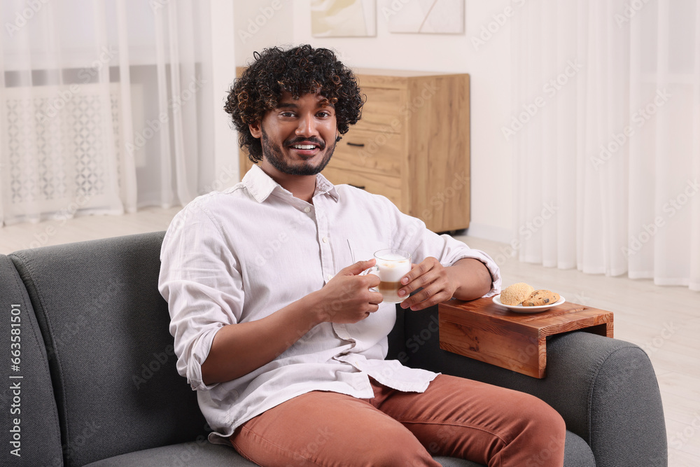 Poster Happy man holding cup of drink at home. Cookies on sofa armrest wooden table