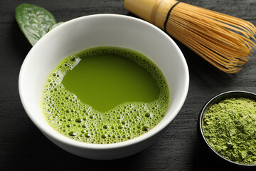 Cup of fresh matcha tea, green powder and bamboo whisk on black wooden table, closeup
