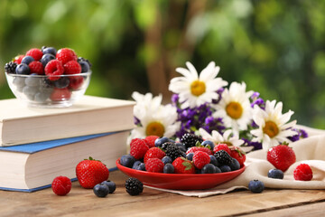 Different fresh ripe berries, beautiful flowers and books on wooden table outdoors