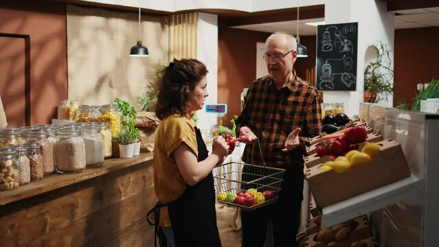 Sustainable zero waste supermarket seller assisting aged client, adding needed food items to shopping basket. Storekeeper helping old man in local shop, teaching him how to prepare bought ingredients