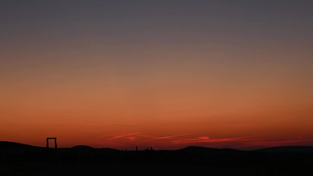 Time lapse of slow moving clouds in an orange sunset colored sky. Old fence posts and rolling hills occur as silhouette shapes along the horizon.    
