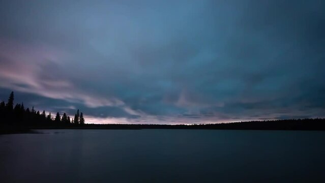 Time lapse of fast-moving night clouds moving towards the viewer over a large lake with a treed shoreline. The water surface is smooth and reflects the color tones from the sky.
