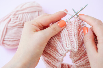Female hands knit a hat with knitting needles on a pink background close-up.