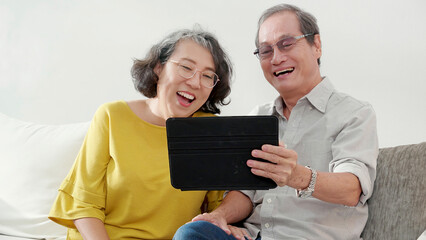 Happiness senior couple sitting on sofa using tablet while video call online with family in living room at home, happy elderly man and woman conversation distant with family with enjoy and relaxation.