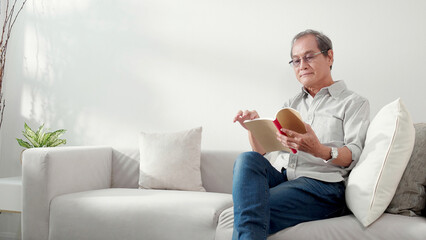 Asian senior man reading a book sitting on sofa with relax in living room at home, elderly man...