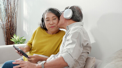 Happy family senior couple wearing headphones listening music and watching tablet to social media while man kiss to cheek woman in living room at home, elderly man and woman relax and cozy with enjoy. - obrazy, fototapety, plakaty