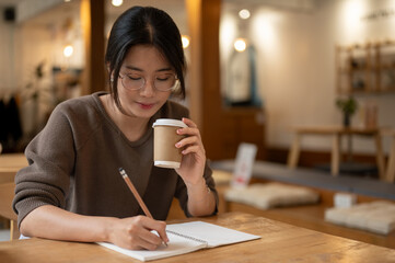A pretty Asian woman focuses on keeping her diary and sipping coffee in a coffee shop.