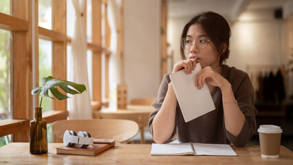 A pensive, thoughtful young Asian woman sits at a table in a beautiful, cosy coffee shop.