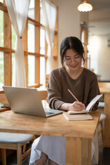 A woman is working remotely at a coffee shop, taking notes in her book and working on her laptop.