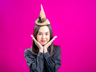 Portrait of an Asian Indonesian woman wearing a Halloween-themed costume with a witch hat, posing with her hands on her chin. Isolated against a magenta background.