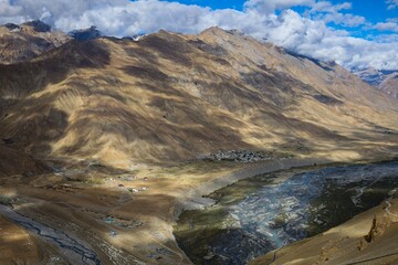 Scenic shot near Kaza, Spiti Valley, India: Brown soils, cloudy shadows, and the stunning blue Spiti River with lush greenery along its shores.