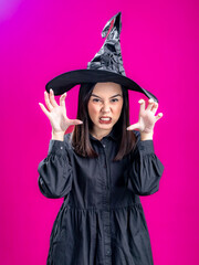 Portrait of an Asian Indonesian woman wearing a Halloween-themed costume with a witch hat, posing in a scary manner with her hands in a claw-like gesture. Isolated against a magenta background.