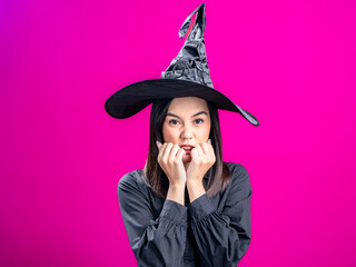 Portrait of an Asian Indonesian woman wearing a Halloween-themed costume with a witch hat, looking scared with a trembling pose. Isolated against a magenta background.