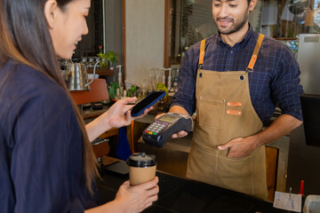 Young Indian baristas or cashier workers man holding a payment machine for scan, cashless technology and credit card payment concept. Customer using smartphone for payment to owner at cafe restaurant