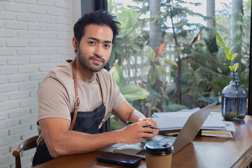 Handsome Indian male coffee shop owner sit and looking at camera with confidence. Man employee...