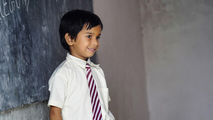 Cute Happy Indian school boy wearing uniform in the classroom near the chalk board