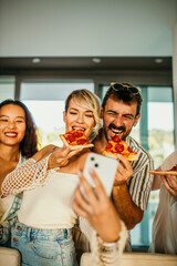 Diverse group of pals enjoying a fun indoor gathering with pizza and drinks, capturing the moment with selfies and laughter