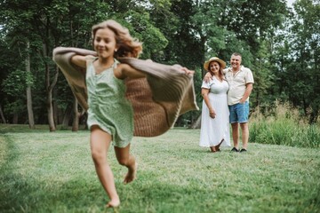 Obraz premium Lovely girl in fluttering knitted cardigan is running across lawn, smiling parents are watching. 