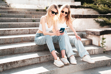 Two young beautiful smiling trendy female. Carefree women posing in street. Positive models having fun. Cheerful and happy. Hold smartphone, look at mobile cellphone screen, use phone apps