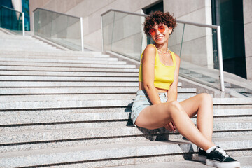 Young beautiful smiling hipster woman in trendy summer clothes. Carefree woman with curls hairstyle, posing on the street background at sunny day. Positive model outdoors. Cheerful and happy