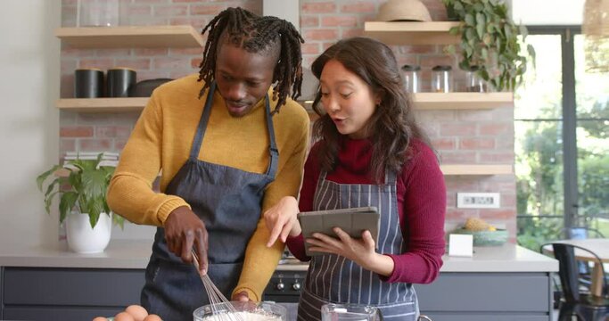 Happy diverse couple in aprons using tablet and baking in sunny kitchen, slow motion