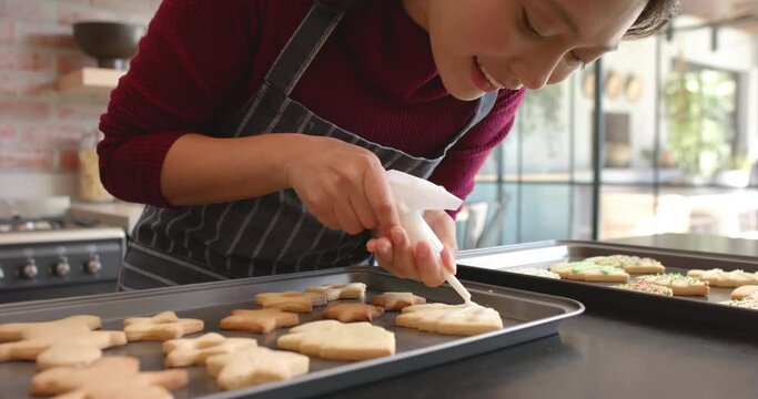 Happy biracial woman in apron decorating christmas cookies in kitchen, slow motion