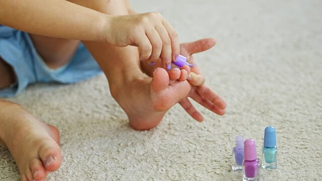 Close up view of hands of little girl doing pedicure and painting nails with colorful pink, blue and purple nail polish at home living room
