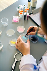 Back view of unrecognizable female chemist examining blue glitter in facial cream sample over petri dish on environment research laboratory. Scientist studying microplastics on cosmetic composition. - obrazy, fototapety, plakaty