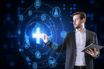 Attractive thoughtful young businessman with tablet using creative glowing blue medical hologram with cross and globe on dark background. Science, healthcare system and futuristic pharmacy concept.