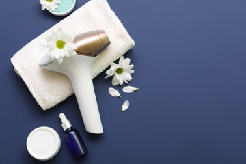 Obraz na płótnie Canvas Modern photoepilator with beautiful flowers, bath towel and cosmetic products on dark blue background