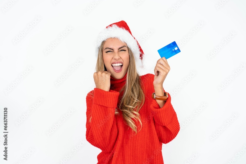 Wall mural Excited happy positive cheerful smiling Beautiful hispanic woman wearing christmas hat and red knitted sweater hold credit card raise fist in victory