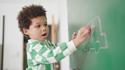 African american cute little child boy writes on a chalk board with chalk in classroom at school. Little boy writes on green board in classroom. Back to School. Education concept - obrazy, fototapety, plakaty