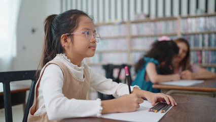 Obraz premium Asian school girl in glasses studying, writing notes in notebook at classroom. Schoolgirl studying and preparing for exams. little pupil writing at desk in classroom. Education knowledge concept